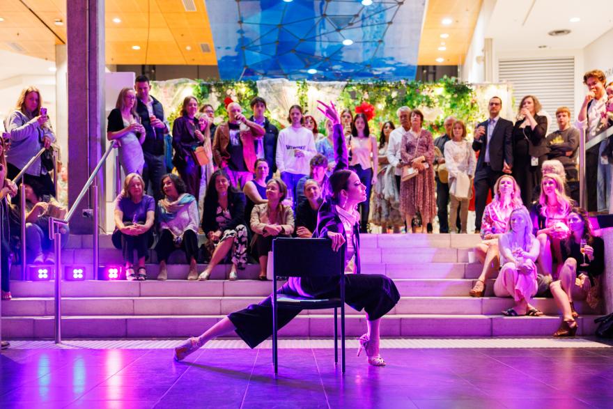 A dancer in black poses on a chair under pink and purple lights, surrounded by an attentive audience on steps, in a modern, vibrant space.