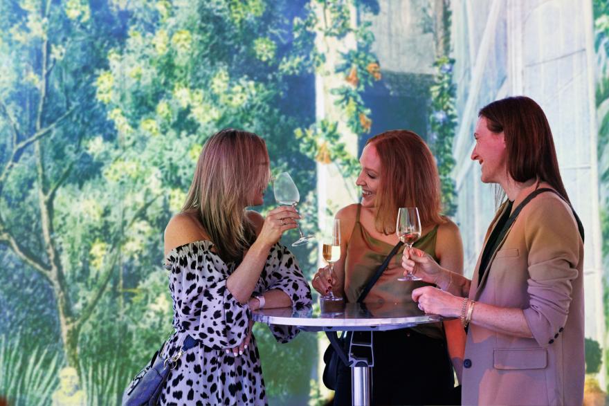 Three women standing, holding wine glasses, against a backdrop of leafy green art.