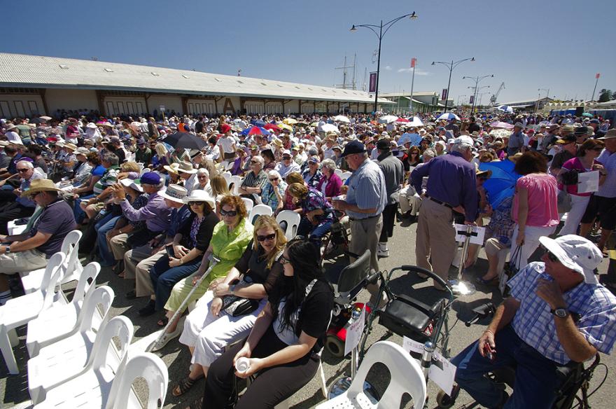 A large crowd of people seated in rows on white plastic chairs. The rows stretch along a large maritime shed