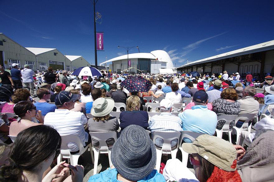 A large crowd of people seated in rows on white plastic chairs with the dome-shaped roof of the Maritime Museum in the background.
