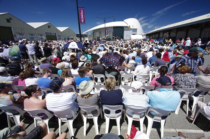 A large crowd of people seated in rows on white plastic chairs with the dome-shaped roof of the Maritime Museum in the background.
