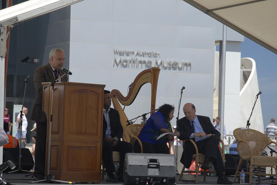 A person in a brown suit stands at a wooden lectern on a stage while three individuals seat beside them. A white building with the words Western Australian Maritime Museum is visible in the background
