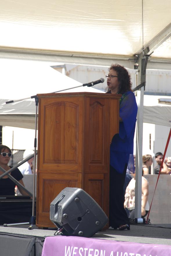 A person with curly brown hair and a long purple shirt stands behind a wooden lectern and speaks into a microphone.