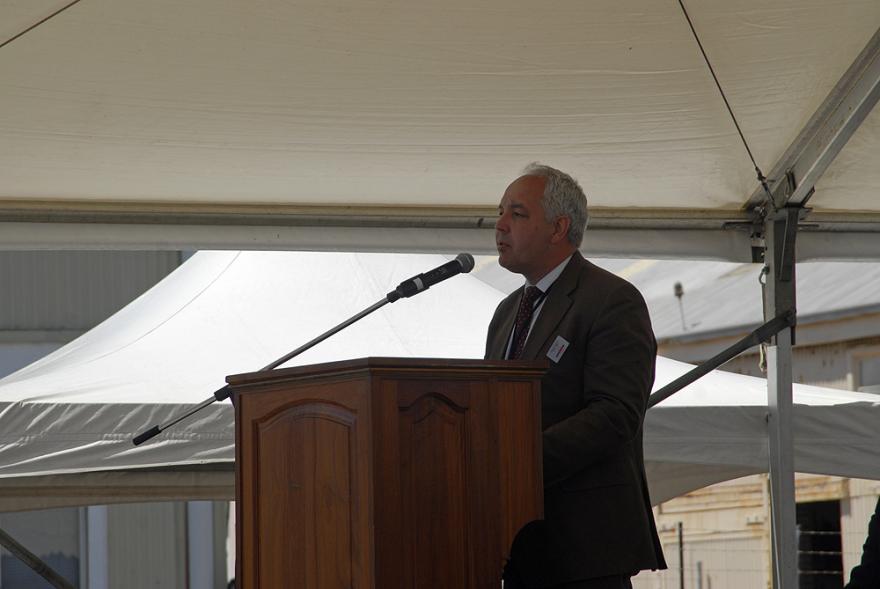 A person wearing a brown suit with white hair stands behind a wooden lectern and speaks into a microphone.