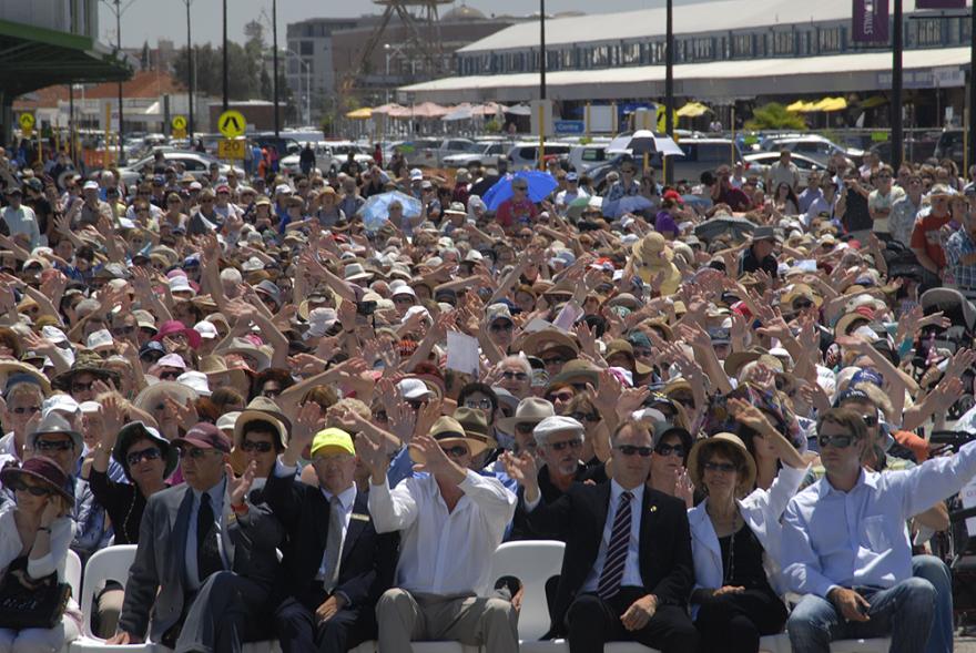 A large crowd of people seated in rows on white plastic chairs and all hold up their hands as though waving at the camera