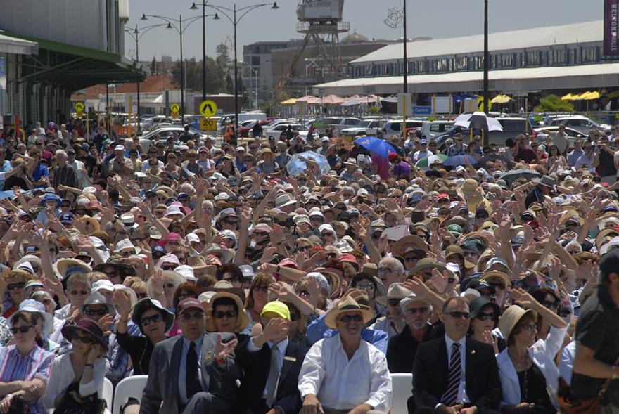 A large crowd of people seated in rows on white plastic chairs and all hold up their hands as though waving at the camera