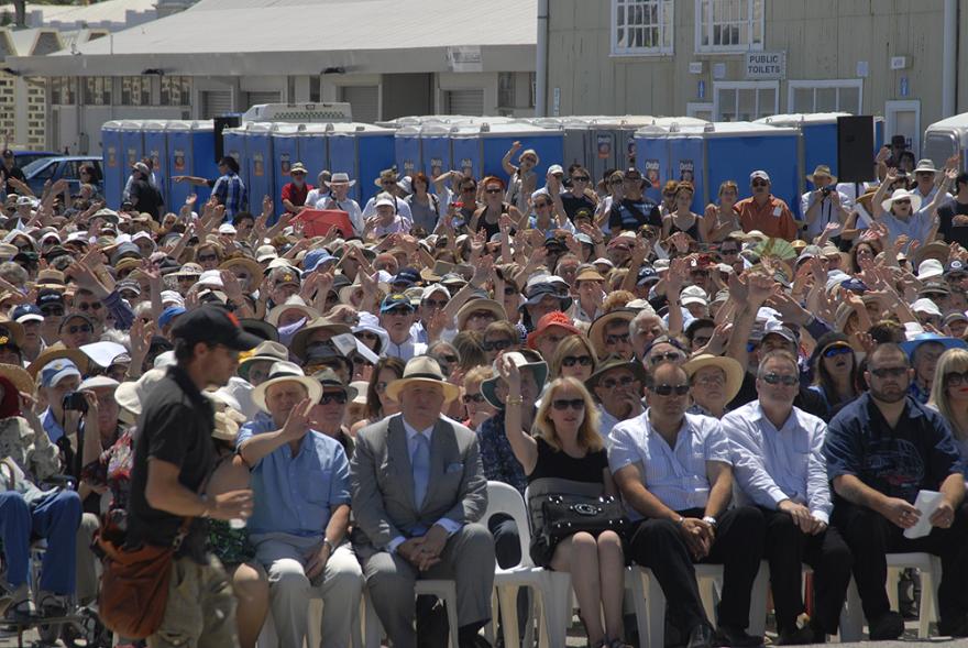 A large crowd of people seated in rows on white plastic chairs and all hold up their hands as though waving at the camera