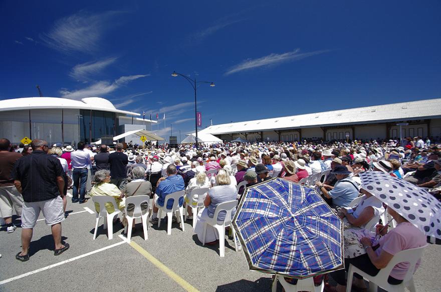A large crowd of people seated in rows on white plastic chairs with the dome-shaped roof of the Maritime Museum in the background. Several patterned umbrellas are visible in the foreground