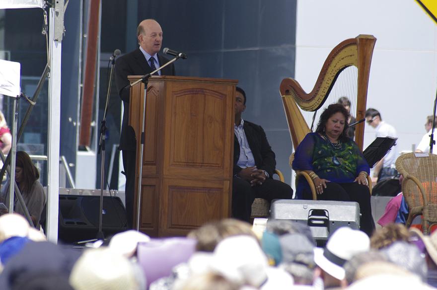 A person in a suit stands at a wooden lectern on a stage next to two images sat on chairs