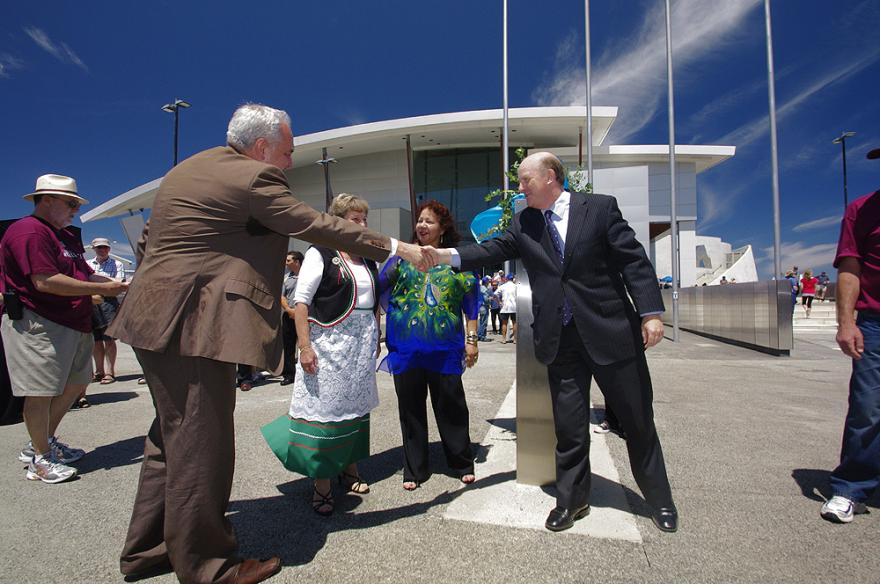 Two people in suits shake hands while a person in a bright blue shirt with a peacock, and someone in cultural dress stand by watching. The curved white roof of the Maritime Museum is visible in the background.