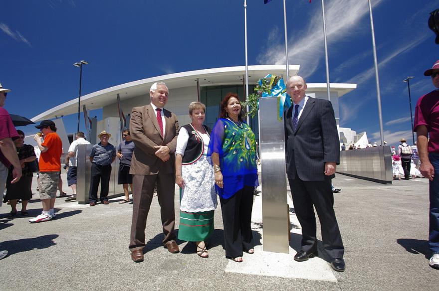 Four individuals in suits and cultural dress pose for a photograph beside a silver installation in front of the large white Maritime Museum building