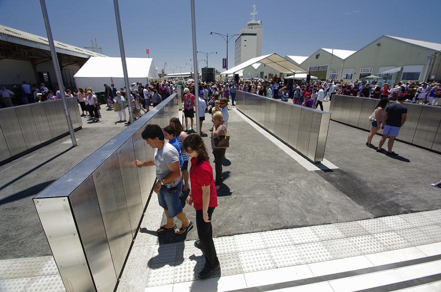 Crowd gathered in an outdoor quay examining metal panels with engravings of names.