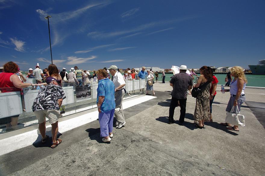 Crowd gathered in an outdoor quay examining glass panels with engravings of names. The ocean is visible in the background.