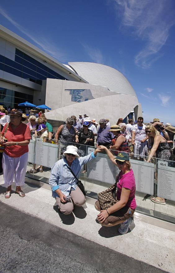 Two individuals crouch down next to a glass wall with engraved names for a photograph with the Maritime Museum visible in the background