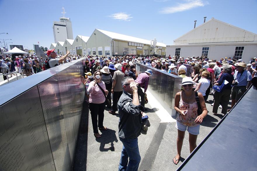 Crowd gathered in an outdoor quay examining metal panels with engravings of names.