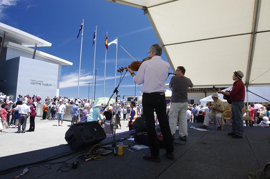 Outdoor event with musicians performing on a stage in front of a Maritime Museum, with a large crowd and flags in the background on a sunny day.