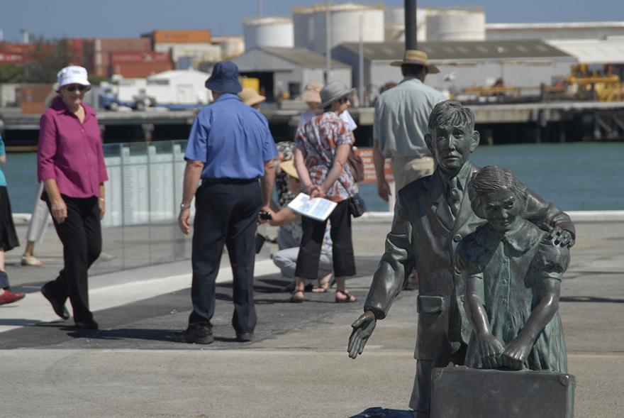 Statue of a boy and girl holding a suitcase at a harbour with people walking nearby.