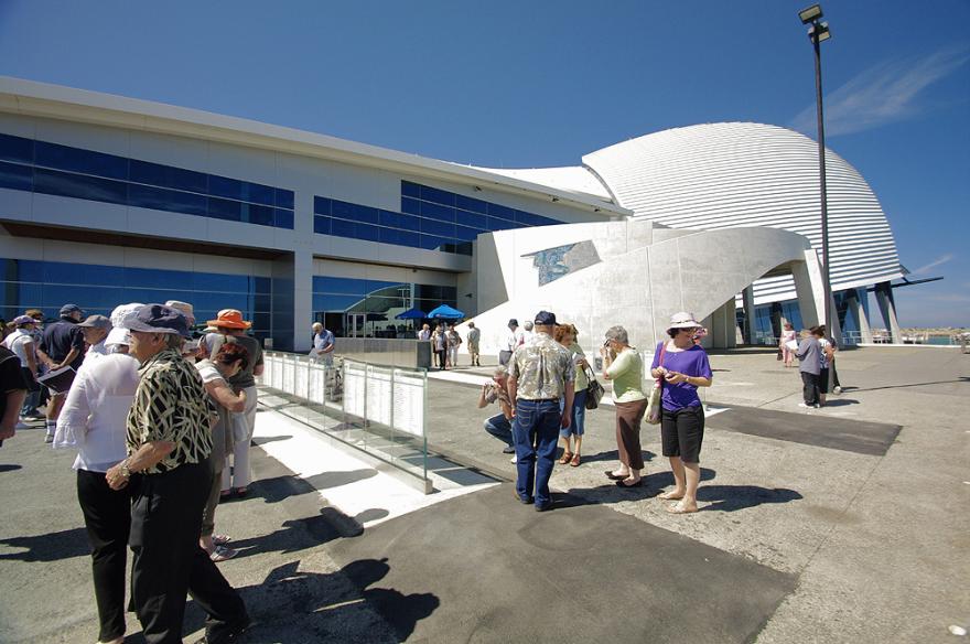 People gathered outside the Maritime Museum which has the appearance of a modern building with blue-tinted windows and a curved white structure.