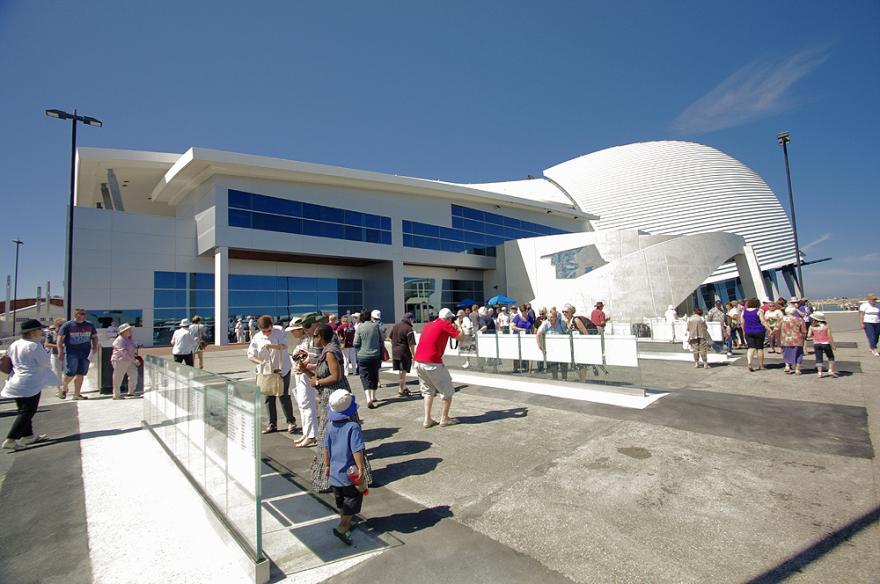 People gathered outside the Maritime Museum which has the appearance of a modern building with blue-tinted windows and a curved white structure.