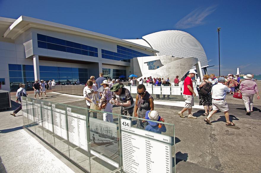 People gathered outside the Maritime Museum which has the appearance of a modern building with blue-tinted windows and a curved white structure. They read from panels of glass with text on it at the front of the Museum