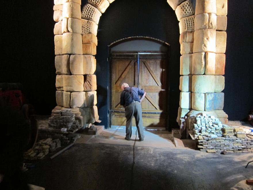 A person in grey trousers and suspenders over a dark blue sweeping in a dimly lit museum gallery under a portico made from large brick slabs. Piles of smaller bricks cover the floor.