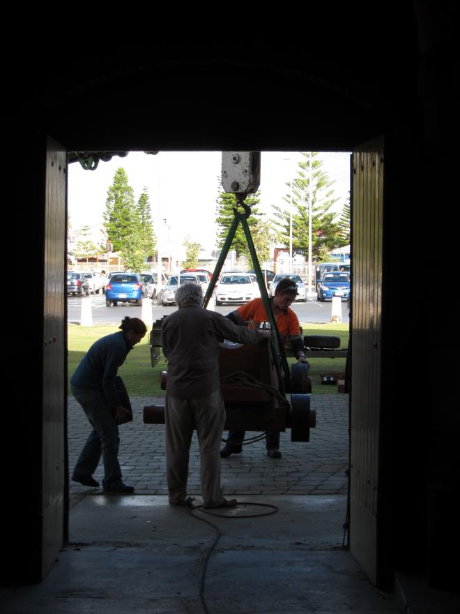 An open doorway into a dark room where three individuals support a large cannon hoisted on a crane arm into the building. A carpark and grass is visible in the distance.