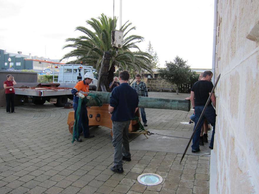People moving a large cannon on a wooden carriage in a paved outdoor setting with a truck and palm trees in the background.