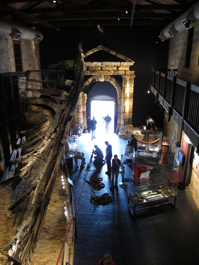 Museum interior with large wooden ship structure, glass display cases, and a stone portico. Individuals work in the space around construction equipment.