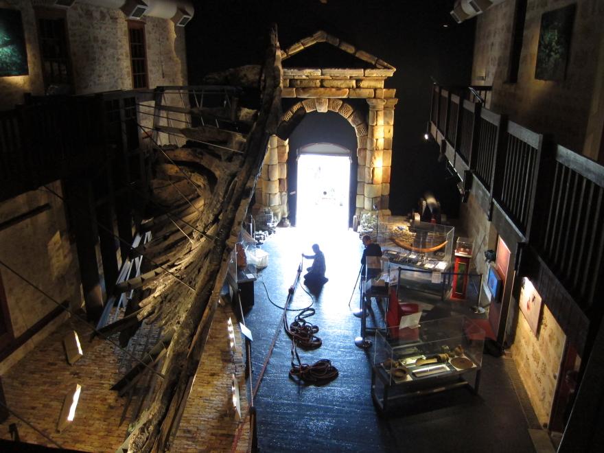 Museum interior with large wooden ship structure, glass display cases, and a stone portico. Individuals work in the space around construction equipment.