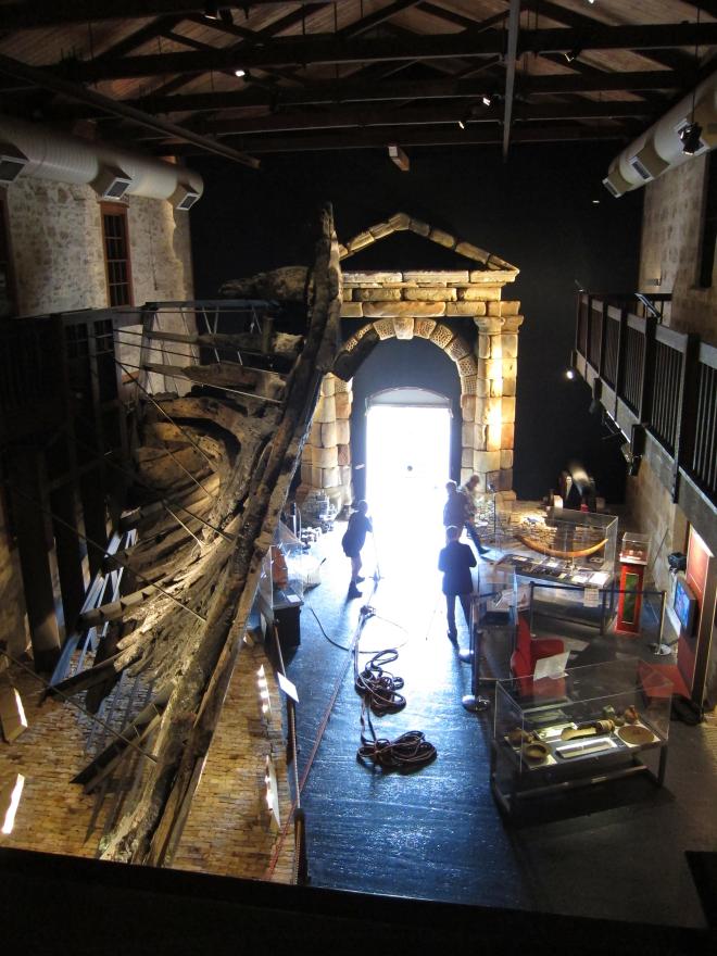 Museum interior with large wooden ship structure, glass display cases, and a stone portico. Individuals work in the space around construction equipment.