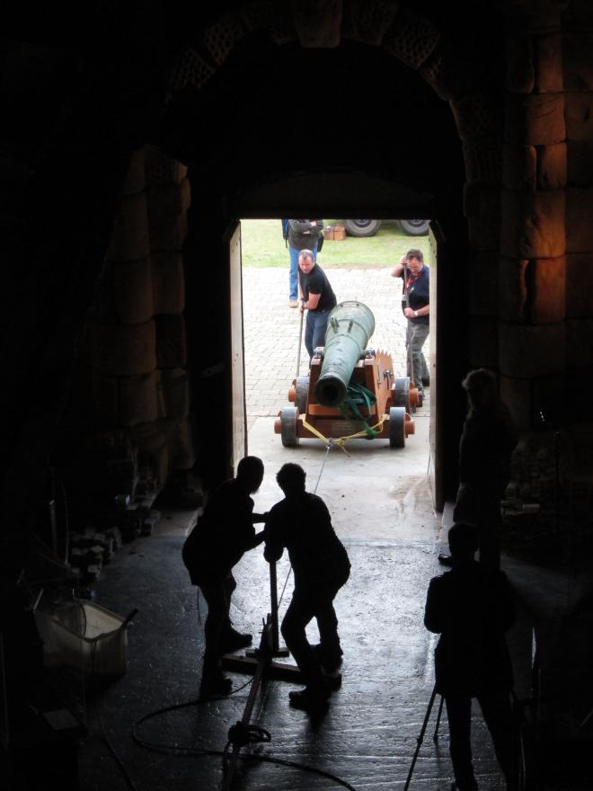 Dark room with a rope leading to a bright open doorway where several people pull and push a large cannon in via a rope system.