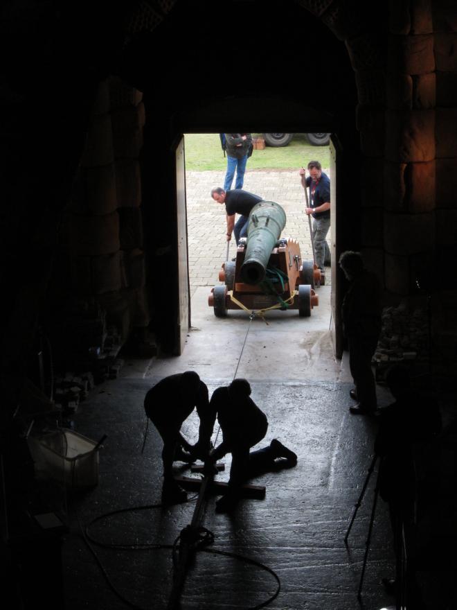 Dark room with a rope leading to a bright open doorway where several people pull and push a large cannon in via a rope system.