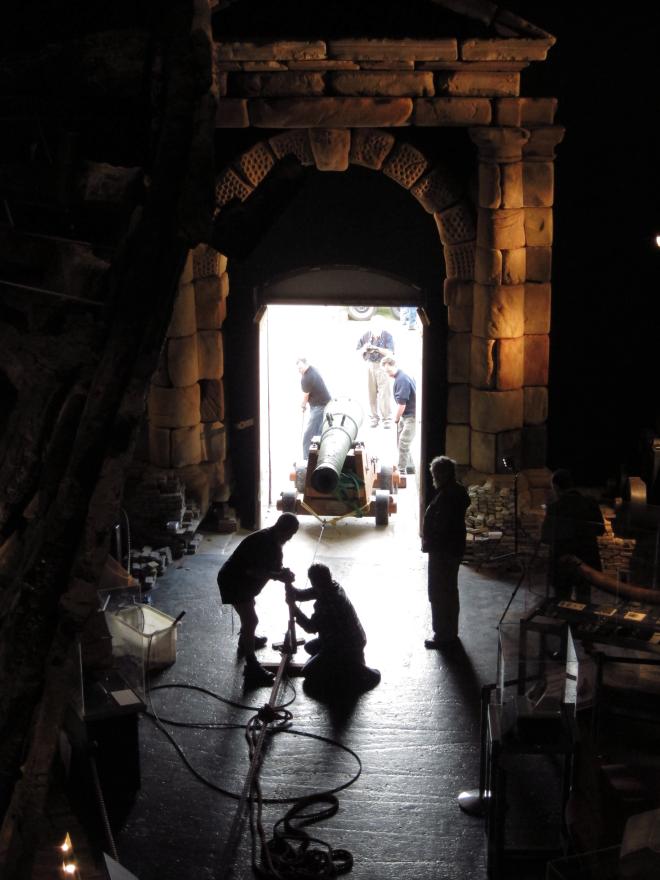 Dark room with a rope leading to a bright open doorway where several people pull and push a large cannon in via a rope system.