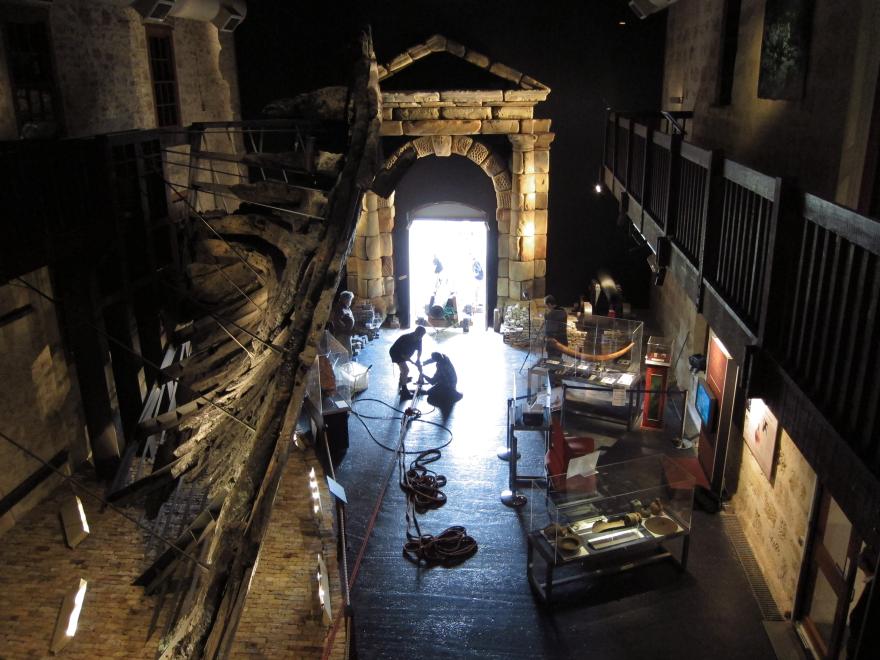 Dark museum room with large wooden ship structure, glass display cases, and a stone portico leading to a bright open doorway where several people pull and push a large cannon in via a rope system.