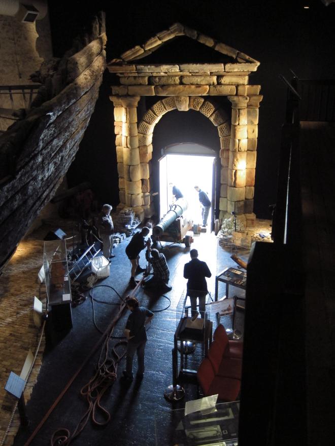 Dark museum room with large wooden ship structure, glass display cases, and a stone portico leading to a bright open doorway where several people pull and push a large cannon in via a rope system.