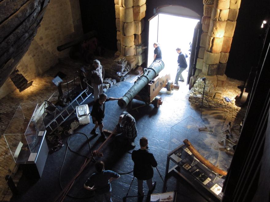 Dark museum room with large wooden ship structure, glass display cases, and a stone portico leading to a bright open doorway where several people pull and push a large cannon in via a rope system.