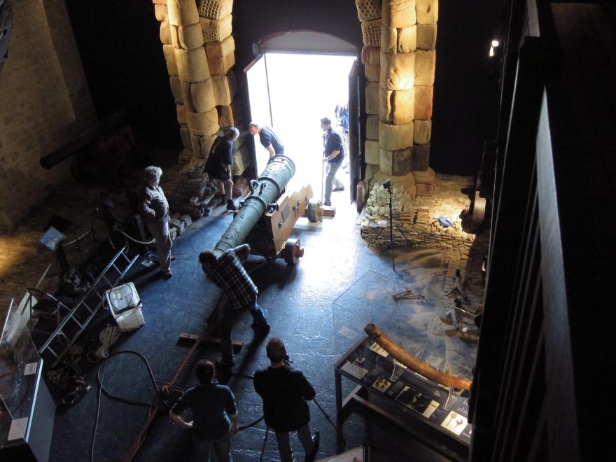 Dark museum room with large wooden ship structure, glass display cases, and a stone portico leading to a bright open doorway where several people pull and push a large cannon in via a rope system.