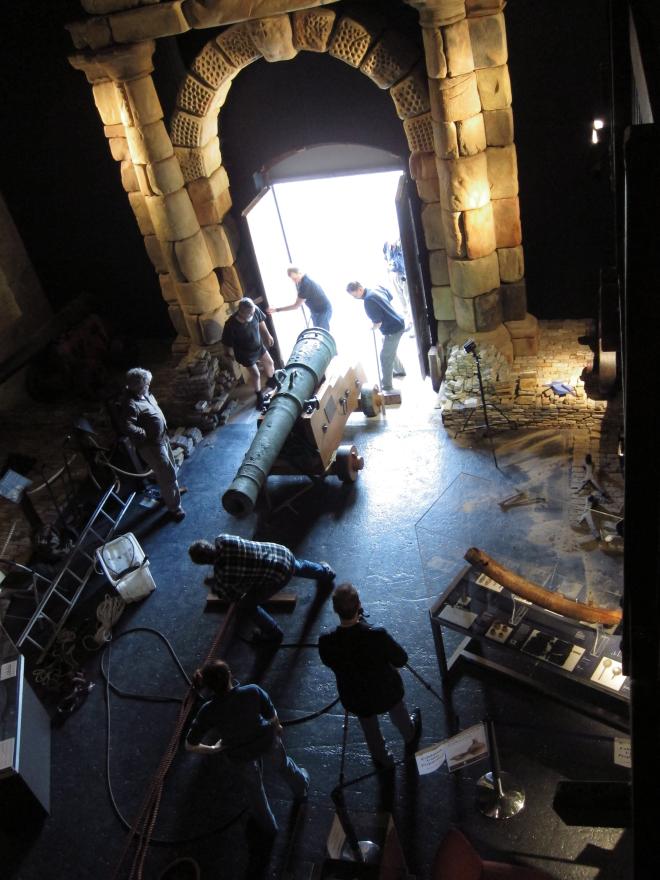 Dark museum room with large wooden ship structure, glass display cases, and a stone portico leading to a bright open doorway where several people pull and push a large cannon in via a rope system.
