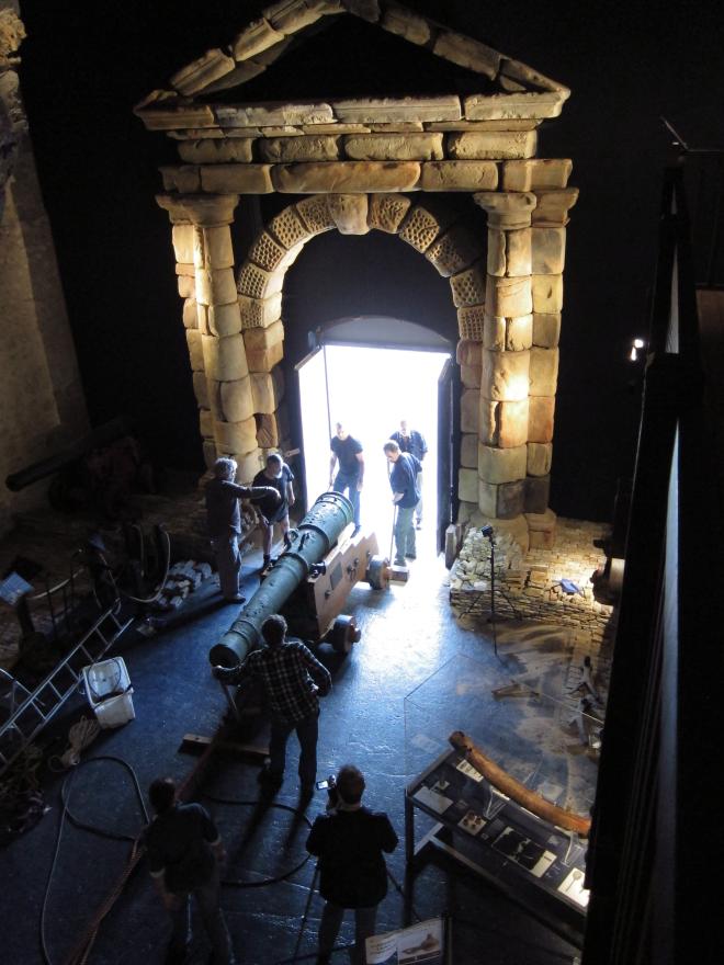 Dark museum room with large wooden ship structure, glass display cases, and a stone portico leading to a bright open doorway where several people pull and push a large cannon in via a rope system.