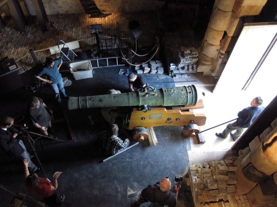 Overhead shot of a large cannon being guided into a dimly lit museum gallery with eight people assisting or overseeing its transportation.