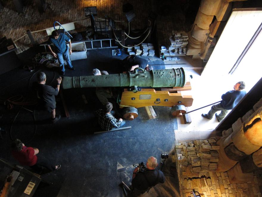 Overhead shot of a large cannon being guided into a dimly lit museum gallery with eight people assisting or overseeing its transportation.