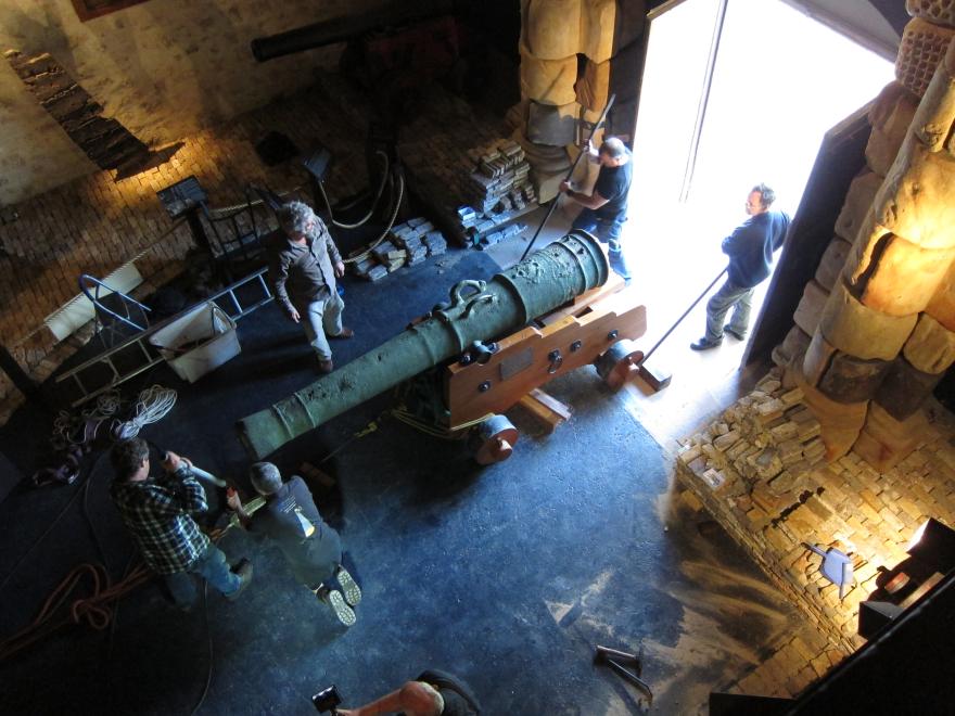 Overhead shot of a large cannon being guided into a dimly lit museum gallery with eight people assisting or overseeing its transportation.