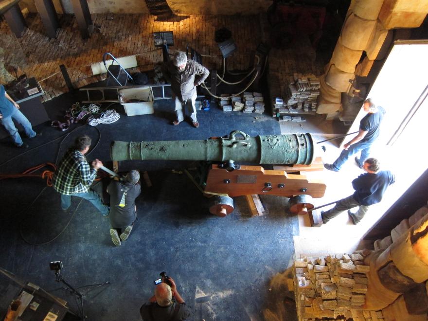 Overhead shot of a large cannon being guided into a dimly lit museum gallery with eight people assisting or overseeing its transportation.