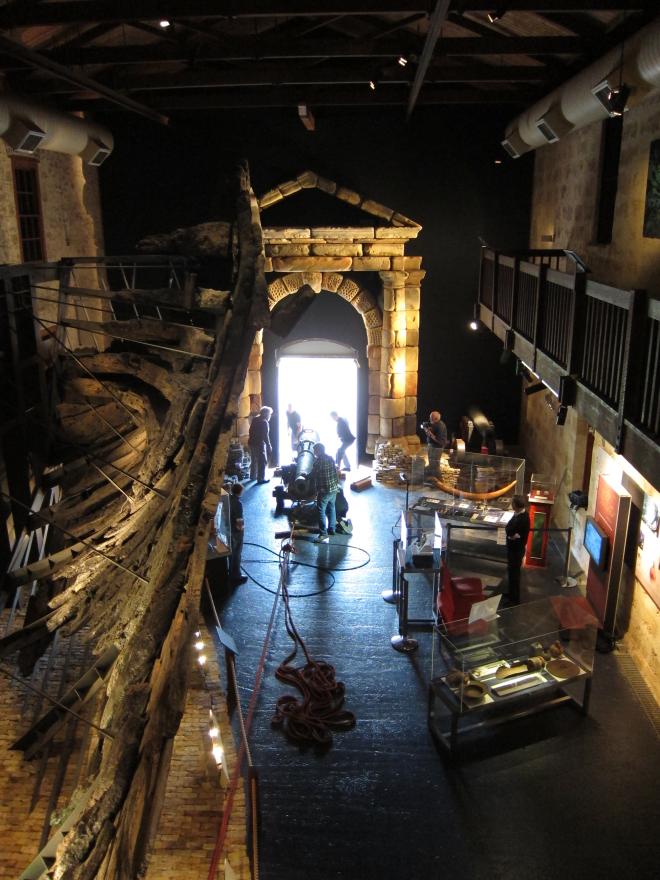 Museum interior with large wooden ship structure, glass display cases, and a stone portico where several people transport in a large cannon in via a rope system.
