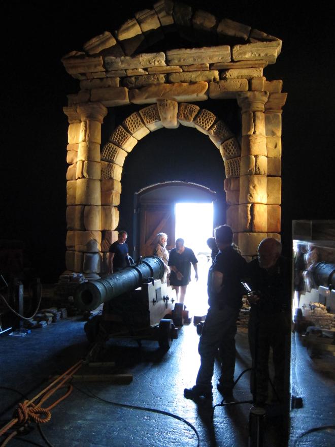 Museum interior with large wooden ship structure, glass display cases, and a stone portico where several people transport in a large cannon in via a rope system.