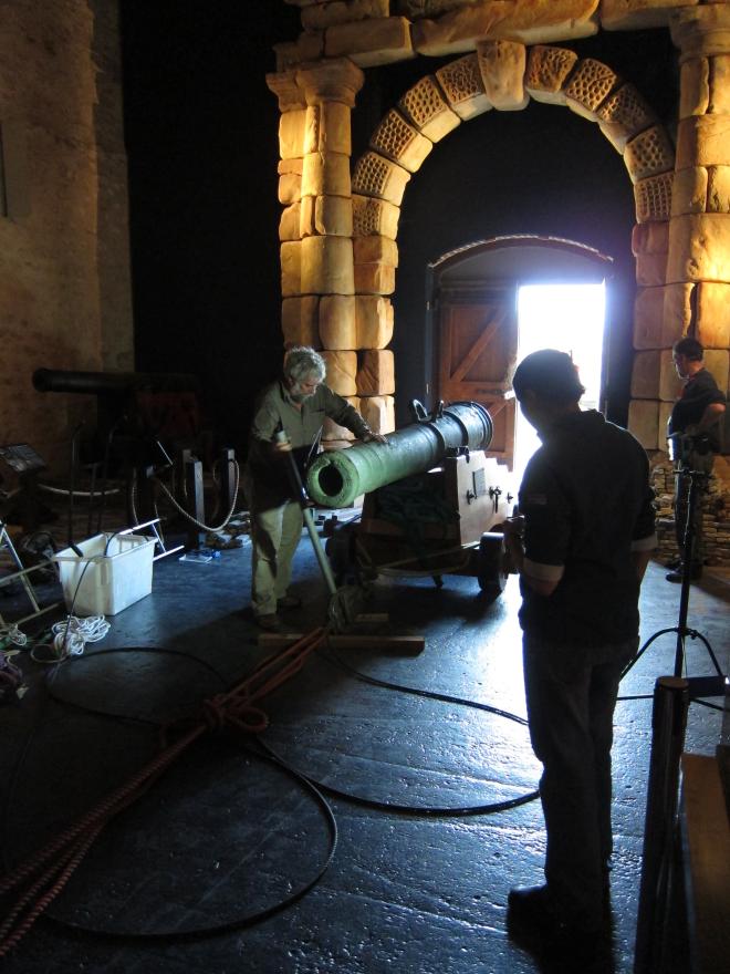 Museum interior with large wooden ship structure, glass display cases, and a stone portico where several people transport in a large cannon in via a rope system.