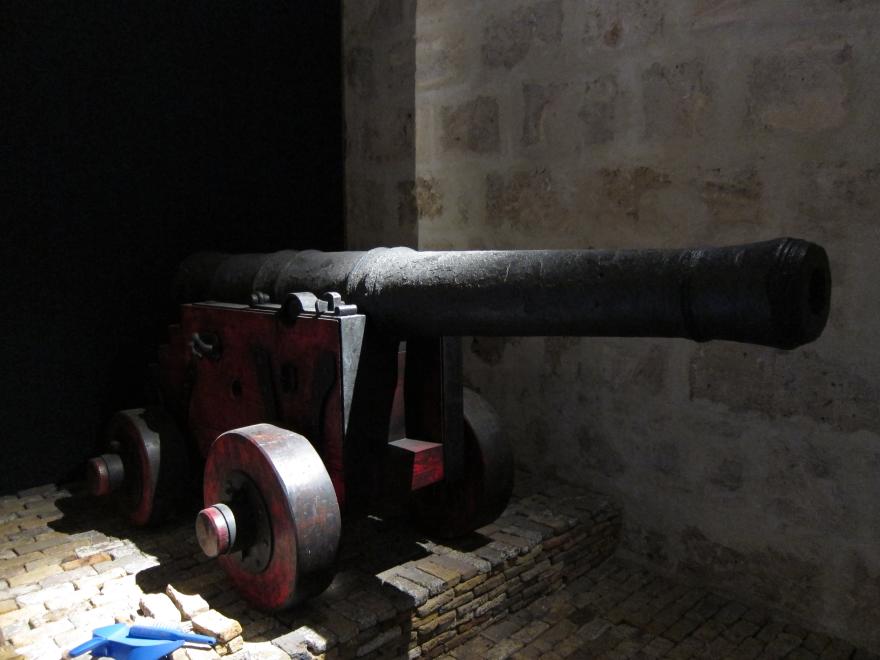 A large canon on a sturdy red base with large round wheels standing on a mount of small worn-looking bricks in a dimly lit museum gallery