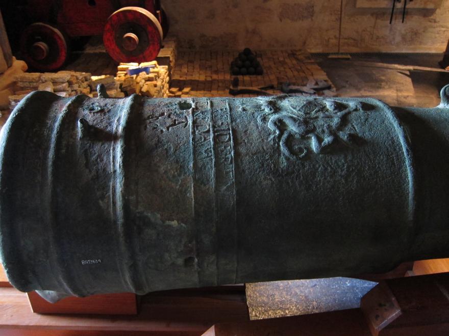 A close up on the end of a large canon showing a series of intricately carved letters and emblems which are partially disfigured by the angle and the canon deterioration.
