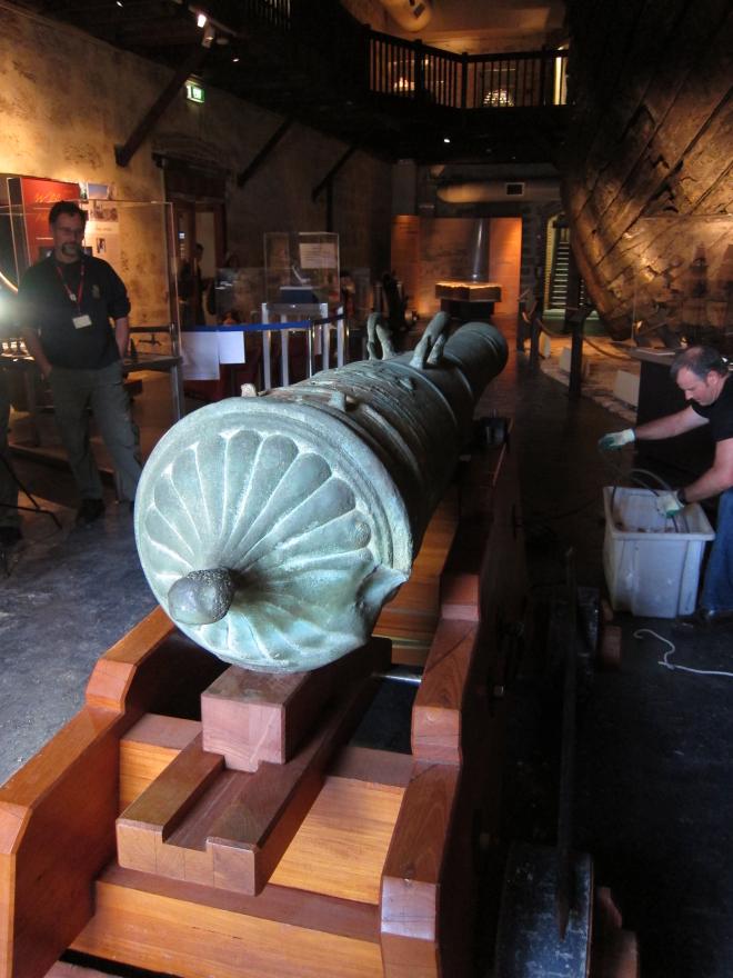An image of the back of a large canon showing its sturdy wooden base and decorative end piece. The canon is situated in a dimly-lit museum gallery.
