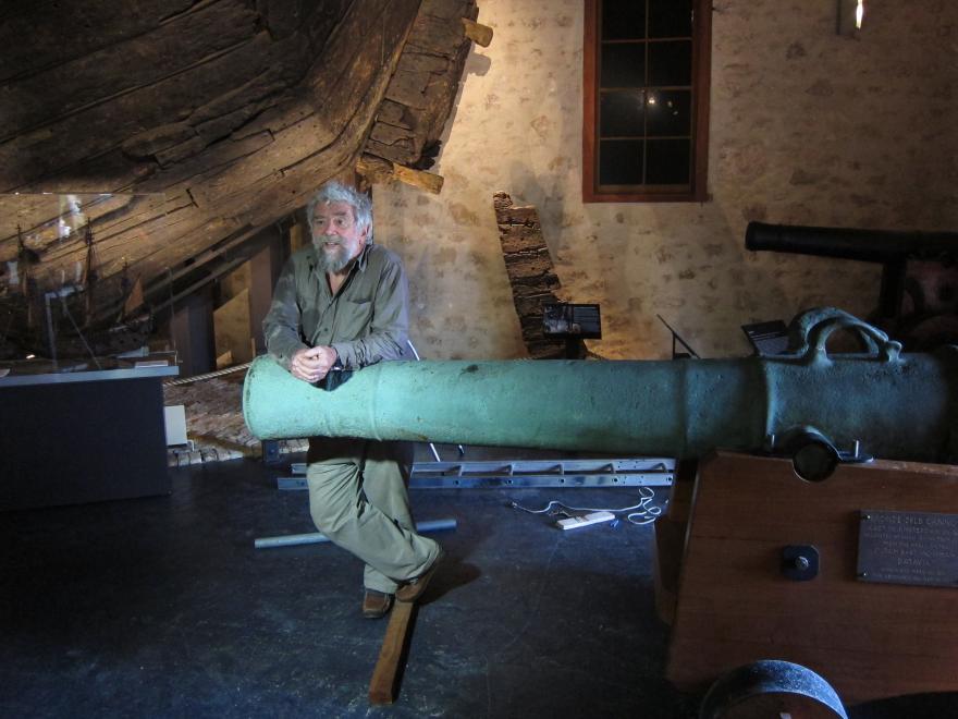 A museum staff member in khaki uniform leans casually on the end of a large canon which appears green in the dim lighting of the museum gallery. A ship is partially visible in the background.